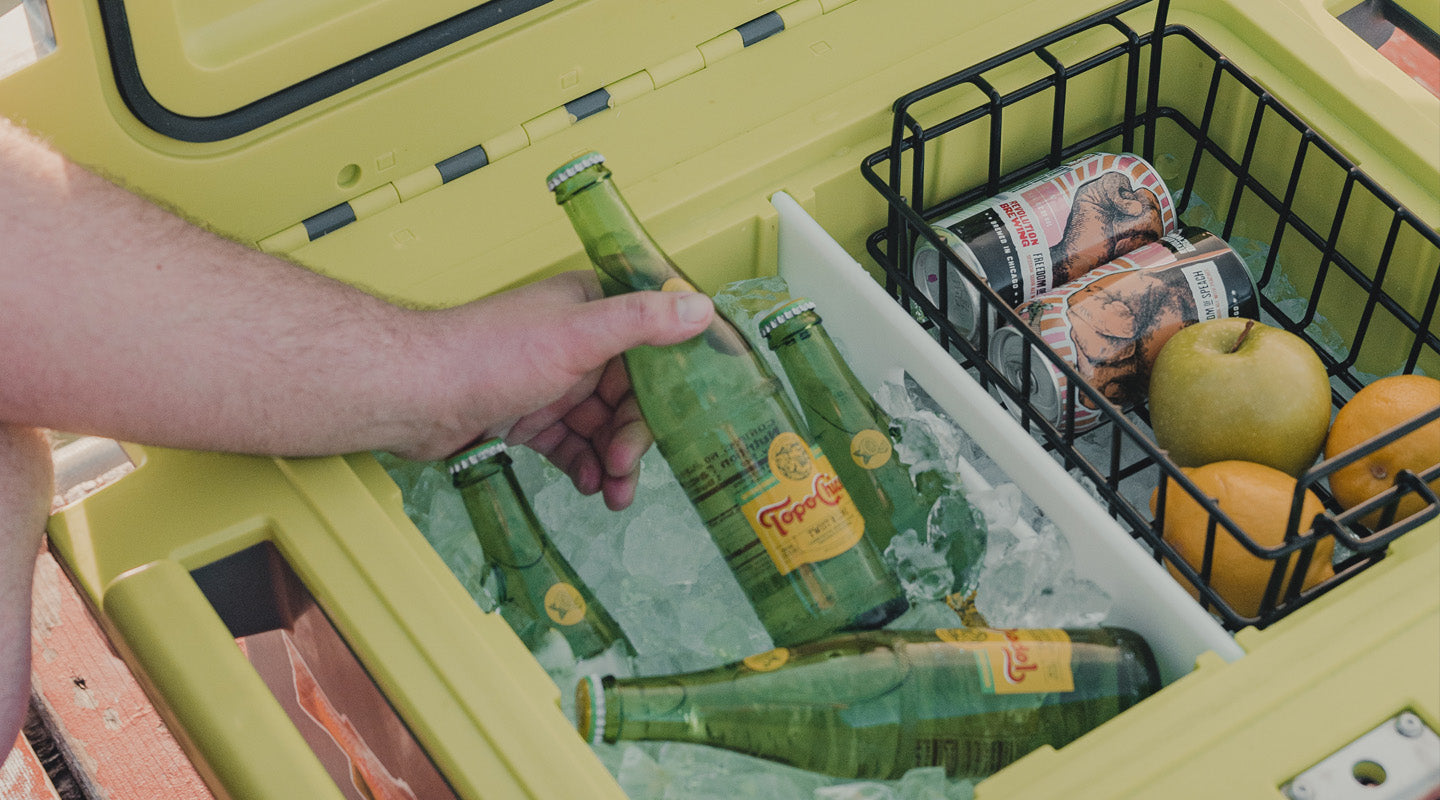 A person is pulling a drink out of a Pelican Elite Cooler. On the right side of the cooler there is a cooler basket filled with fresh fruit and drinks.