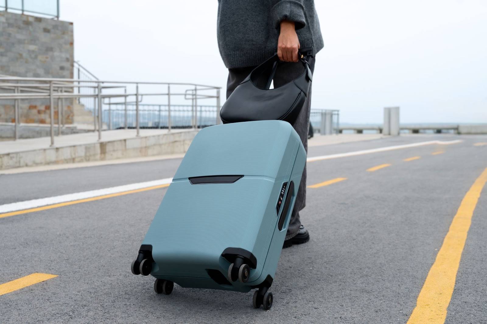 A person is walking on a paved road, pulling a light blue hard shell luggage with a black handbag