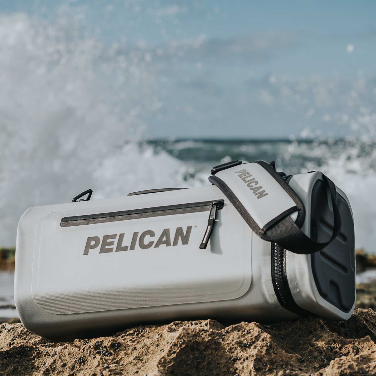 Light Grey Pelican™ Dayventure Sling Soft Cooler on the beach with waves crashing behind it